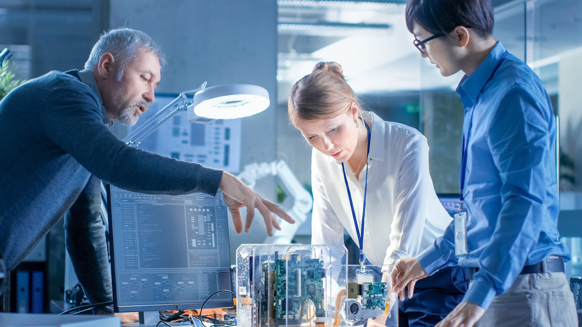 An employee explains a machine part on a table to two colleagues